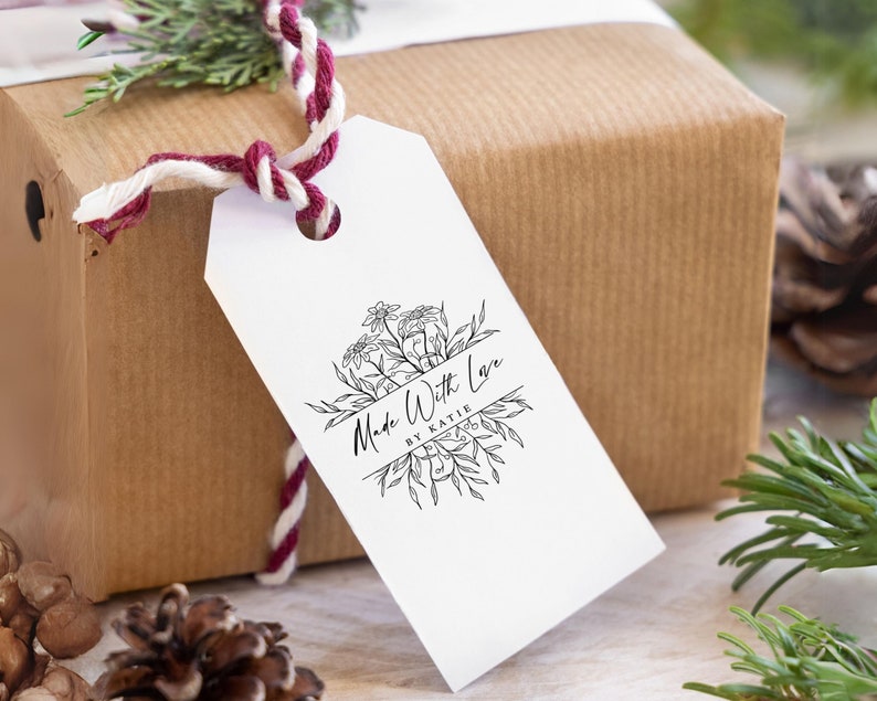 May include: A white rectangular gift tag with a hole at the top, featuring a black ink botanical illustration and the text "Made With Love BY KATIE" in the center. The tag is attached with a twisted red and white cord to a brown paper package.