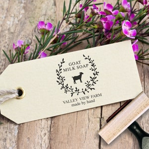 May include: A rectangular beige paper tag with a brass eyelet and jute string, featuring a black ink stamp of a goat silhouette inside a leafy wreath. The text reads GOAT MILK SOAP, VALLEY VIEW FARM, and made by hand. A wooden rubber stamp is nearby.