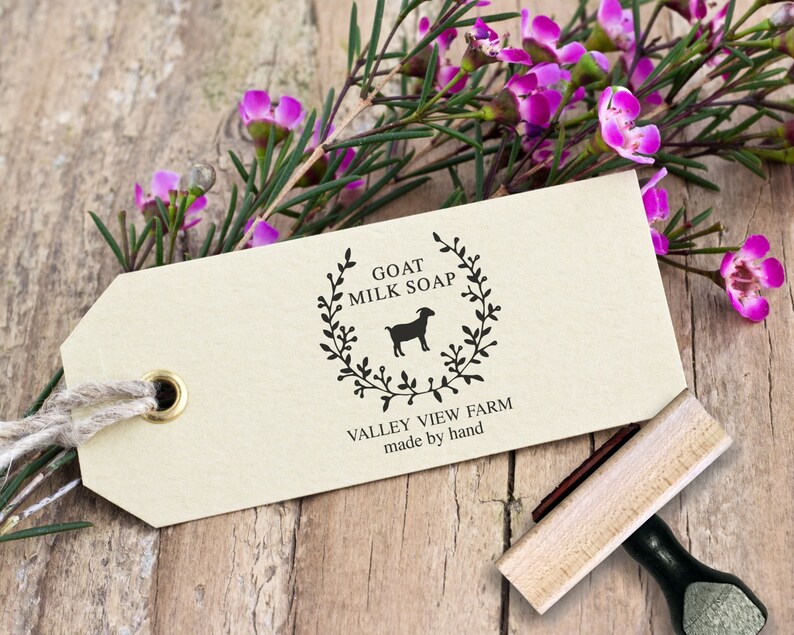 May include: A rectangular beige paper tag with a brass eyelet and jute string, featuring a black ink stamp of a goat silhouette inside a leafy wreath. The text reads GOAT MILK SOAP, VALLEY VIEW FARM, and made by hand. A wooden rubber stamp is nearby.