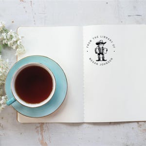 May include: A white notebook with a black and white stamp that says "From the Library of Megan Johnson". The stamp features a cartoon illustration of a person wearing a hat and holding a book. A blue teacup and saucer are on the left side of the notebook.
