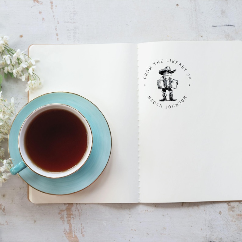 May include: A white notebook with a black and white stamp that says "From the Library of Megan Johnson". The stamp features a cartoon illustration of a person wearing a hat and holding a book. A blue teacup and saucer are on the left side of the notebook.