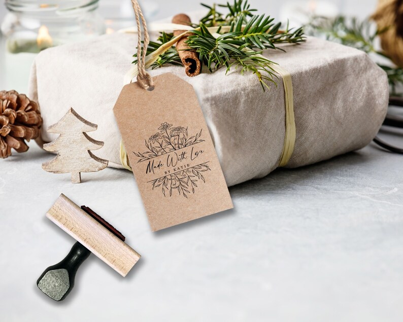 May include: A rectangular brown kraft paper gift tag featuring a black ink botanical illustration and the text Made With Love BY KATIE. The tag is suspended by a jute twine string and is positioned near a wooden stamp with a black handle.