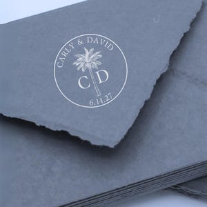 May include: A stack of dark gray envelopes with rough, deckled edges. A circular white stamp is centered on the top envelope, featuring a palm tree graphic, the names CARLY & DAVID, the initials C D, and the date 6.14.27 printed in a clean font.