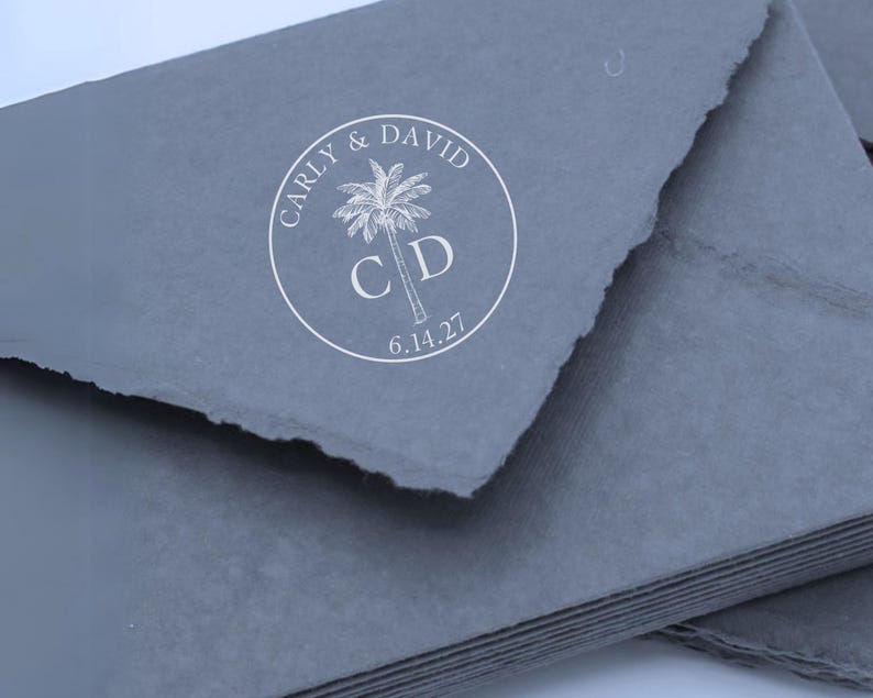May include: A stack of dark gray envelopes with rough, deckled edges. A circular white stamp is centered on the top envelope, featuring a palm tree graphic, the names CARLY & DAVID, the initials C D, and the date 6.14.27 printed in a clean font.