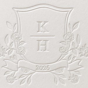 May include: A white embossed card featuring a central shield design with the letters K and H separated by a horizontal line. The shield is surrounded by floral rose motifs and a ribbon at the bottom containing the year 2025 in embossed text.