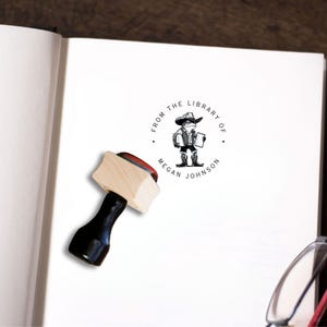 May include: A wooden stamp with a black handle. The stamp features a circular design with the text "From the Library of Megan Johnson" and a cartoon illustration of a frog wearing a cowboy hat and carrying a book.