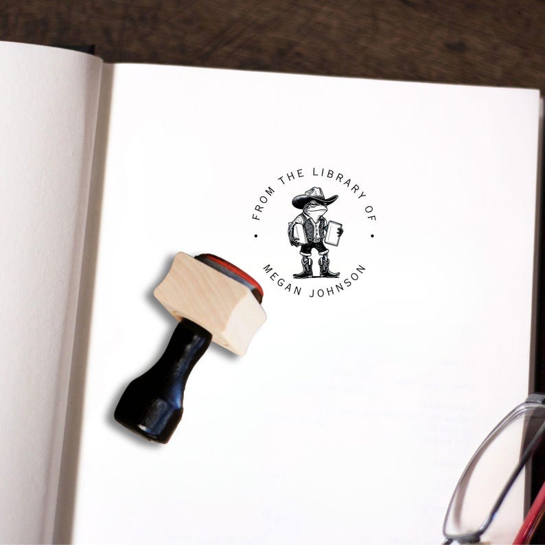 May include: A wooden stamp with a black handle. The stamp features a circular design with the text "From the Library of Megan Johnson" and a cartoon illustration of a frog wearing a cowboy hat and carrying a book.