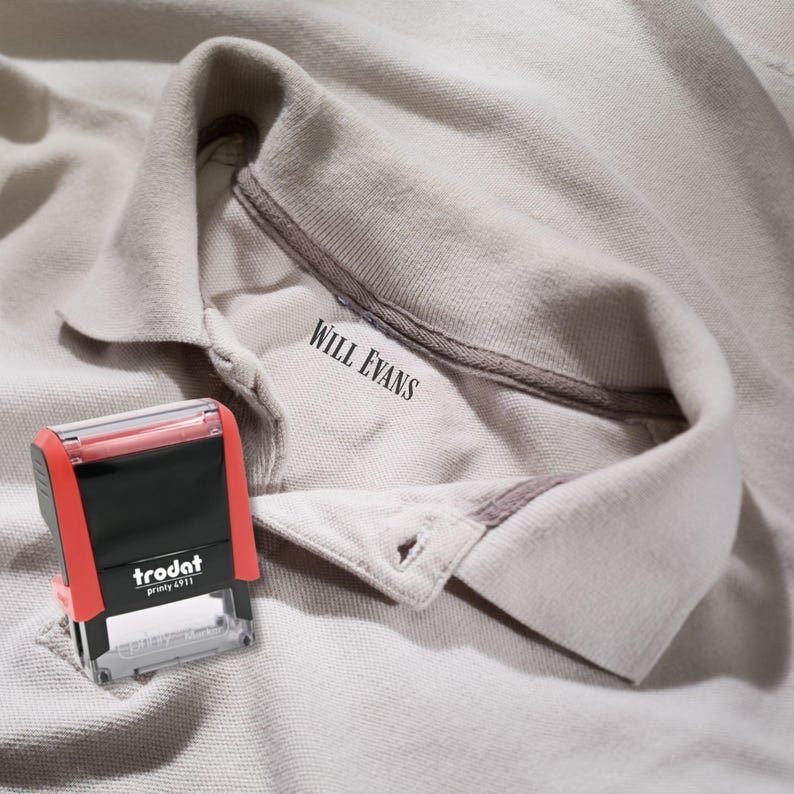 May include: A red and black Trodat 4911 stamp beside a light grey polo shirt. The shirt collar displays the name "WILL EVANS" in black text. The stamp is positioned near the shirt's collar.