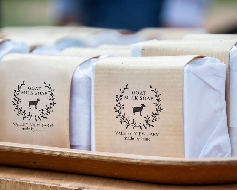 May include: Rectangular bars of soap wrapped in white paper and secured with a tan paper band. The band features a black printed logo with a goat silhouette, a leaf wreath, and the text GOAT MILK SOAP VALLEY VIEW FARM made by hand.