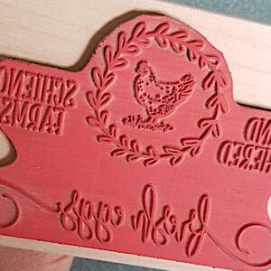 Peut inclure: Un tampon en caoutchouc rouge mont&eacute; sur un bloc de bois clair avec une illustration centrale de poule dans une couronne de feuilles. Le texte indique SCHIENO FARMS, HAND GATHERED et fresh eggs dans un m&eacute;lange de styles d'&eacute;criture.