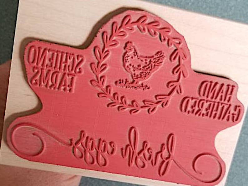 Peut inclure: Un tampon en caoutchouc rouge mont&eacute; sur un bloc de bois clair avec une illustration centrale de poule dans une couronne de feuilles. Le texte indique SCHIENO FARMS, HAND GATHERED et fresh eggs dans un m&eacute;lange de styles d'&eacute;criture.