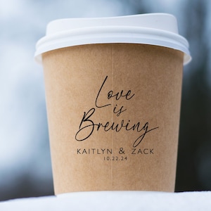May include: A brown paper coffee cup with a white lid. The cup has black text that reads "Love is Brewing" and "Kaitlyn & Zack 10.22.24".