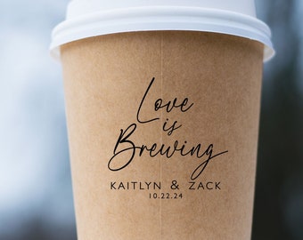 May include: A tan paper coffee cup with a white plastic lid. The cup features black printed text that reads Love is Brewing, followed by the names KAITLYN & ZACK and the date 10.22.24 in a clean, sans-serif font centered on the front surface.