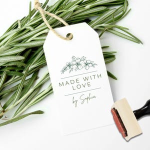 May include: A white rectangular paper tag with a hole at the top and a jute twine loop. The tag features a dark green floral line drawing, the text "MADE WITH LOVE" in capital letters, and the script text "by Sophia" above a rubber stamp.