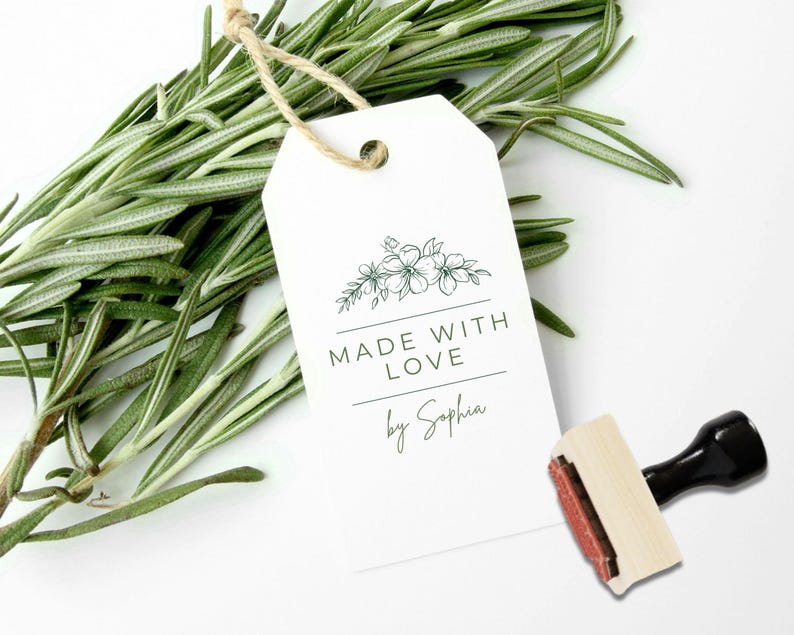 May include: A white rectangular paper tag with a hole at the top and a jute twine loop. The tag features a dark green floral line drawing, the text "MADE WITH LOVE" in capital letters, and the script text "by Sophia" above a rubber stamp.