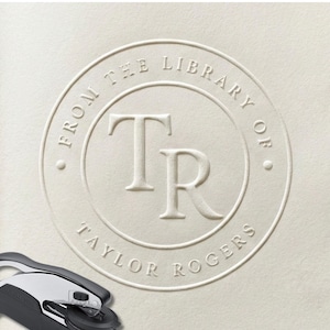 May include: A circular embossed seal on off-white paper featuring the text FROM THE LIBRARY OF and TAYLOR ROGERS surrounding the large central initials TR. A black and silver metal handheld embosser tool is positioned at the bottom left corner.
