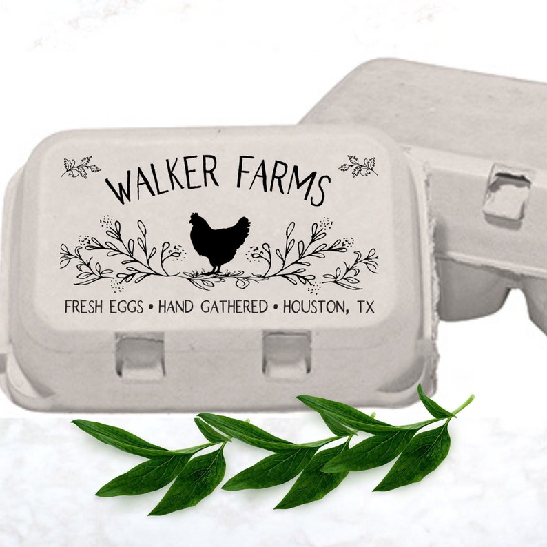 May include: Close-up view of two light gray cardboard egg cartons.  The carton in the foreground features a black and white design with the text 'WALKER FARMS', 'FRESH EGGS', 'HAND GATHERED', and 'HOUSTON, TX'. A black silhouette of a chicken is centered within a decorative floral branch design. A sprig of green leaves rests in front of the cartons.