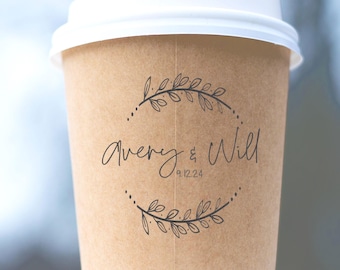 May include: A tan paper coffee cup with a white plastic lid. The cup features a black printed design of two curved leafy branches forming a circle around the text Avery & Will and the date 9.12.24 in a simple, handwritten-style font.