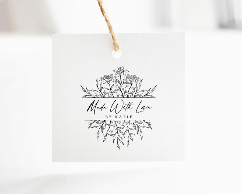 May include: A square white paper tag hanging from a brown twine string. The tag features a black line art illustration of flowers and leaves with the text Made With Love and BY KATIE printed in the center of the floral design.