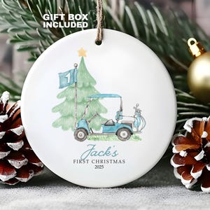 May include: White ceramic Christmas ornament featuring a golf cart, a Christmas tree, and the text "Jack's FIRST CHRISTMAS 2025." The ornament has a string for hanging and is surrounded by pine cones and greenery.