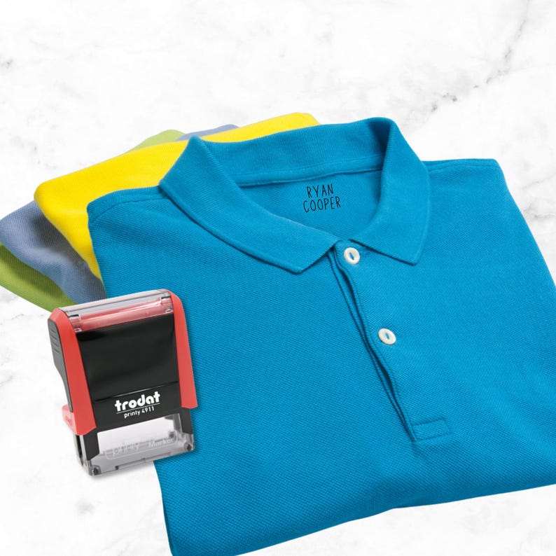 May include: A selection of polo shirts in blue, yellow, and green. A red and black Trodat printy 4911 stamp is in the foreground. The blue shirt has the name "RYAN COOPER" printed on the collar.