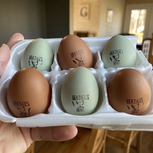 May include: A carton of six eggs in a white foam container. The eggs are various shades of brown and green. Each egg is stamped with the text "Heather's Hens" and a hen graphic.