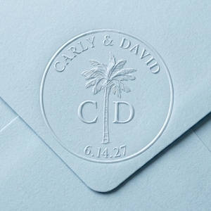 May include: A light blue envelope featuring a circular embossed seal. The design includes a palm tree graphic centered between the letters C and D, with the text CARLY & DAVID arched above and the date 6.14.27 positioned below the palm tree.