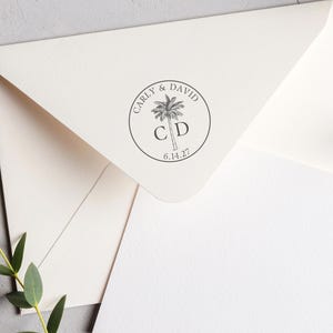 May include: A cream-colored envelope featuring a circular black ink stamp on the flap. The stamp displays a palm tree illustration, the names Carly & David, the initials C D, and the date 6.14.27, all enclosed within a thin circular border.