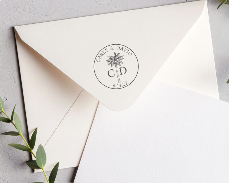 May include: A cream-colored envelope featuring a circular black ink stamp on the flap. The stamp displays a palm tree illustration, the names Carly & David, the initials C D, and the date 6.14.27, all enclosed within a thin circular border.