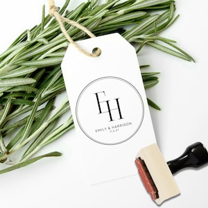 May include: A white tag with a black circular design featuring the initials "EH" and the names "Emily & Harrison" with the date "11.4.27". A wooden stamp with a black handle is next to the tag. Fresh rosemary sprigs are in the background.