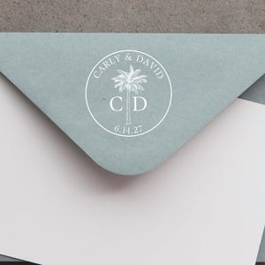 May include: A sage green envelope with a white circular seal featuring a palm tree illustration, the names Carly & David, the initials C D, and the date 6.14.27. A plain white card is tucked inside the envelope flap, partially obscuring the bottom.