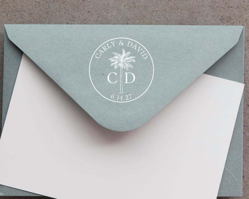 May include: A sage green envelope with a white circular seal featuring a palm tree illustration, the names Carly & David, the initials C D, and the date 6.14.27. A plain white card is tucked inside the envelope flap, partially obscuring the bottom.