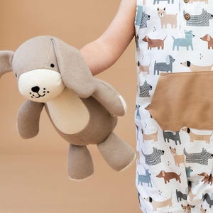 May include: A soft, plush dog toy in shades of brown and cream is held in front of a white onesie patterned with various dog breeds. The onesie has a brown pocket and button details.
