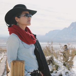 May include: A woman wearing a black cowboy hat, sunglasses, a red scarf, a light blue denim shirt, a black leather jacket, and black jeans. She is standing next to a wooden fence post in a snowy landscape.