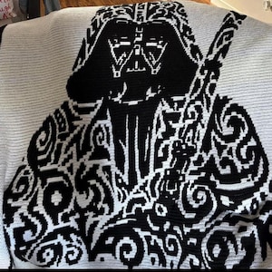 Puede incluir: Una manta de ganchillo en blanco y negro con un diseño de Darth Vader. La manta muestra una imagen grande y pixelada del personaje sobre un fondo blanco. El diseño incorpora intrincados patrones de remolino.