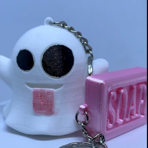 Pode incluir: Um chaveiro de fantasma branco com olhos pretos e uma l&iacute;ngua rosa, preso a um chaveiro de sabonete rosa com a palavra "SOAP" em relevo.