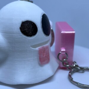 Pode incluir: Um chaveiro fantasma branco com olhos pretos e uma l&iacute;ngua rosa, preso a um chaveiro rosa com um chaveiro prateado.