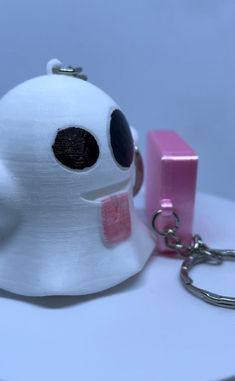 Pode incluir: Um chaveiro fantasma branco com olhos pretos e uma l&iacute;ngua rosa, preso a um chaveiro rosa com um chaveiro prateado.