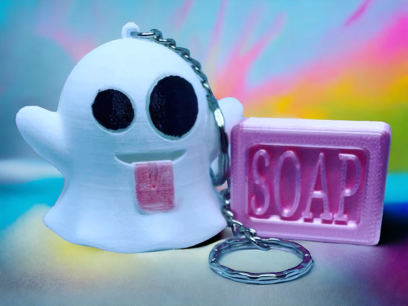 Pode incluir: Um chaveiro fantasma branco com uma l&iacute;ngua rosa e olhos pretos, preso a uma corrente prateada e um chaveiro de sabonete rosa com a palavra "SOAP" impressa.