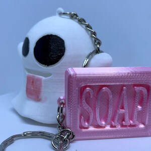 Pode incluir: Um chaveiro fantasma branco com olhos pretos e um chaveiro de sabonete rosa com a palavra "SOAP" em relevo. Os chaveiros s&atilde;o conectados por uma corrente prateada e um anel de chave prateado.