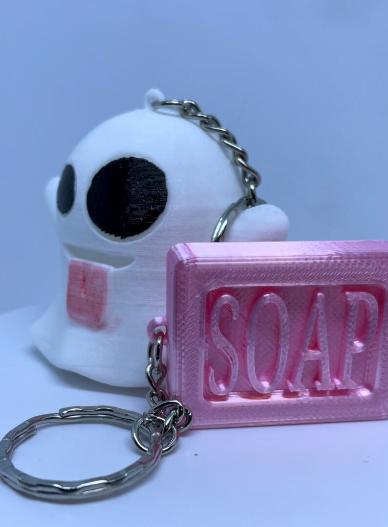Pode incluir: Um chaveiro fantasma branco com olhos pretos e um chaveiro de sabonete rosa com a palavra "SOAP" em relevo. Os chaveiros s&atilde;o conectados por uma corrente prateada e um anel de chave prateado.