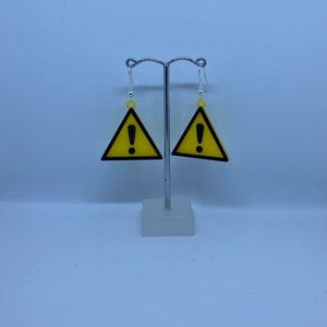 May include: A pair of yellow triangle earrings with a black exclamation point inside. The earrings are hanging from a silver stand.