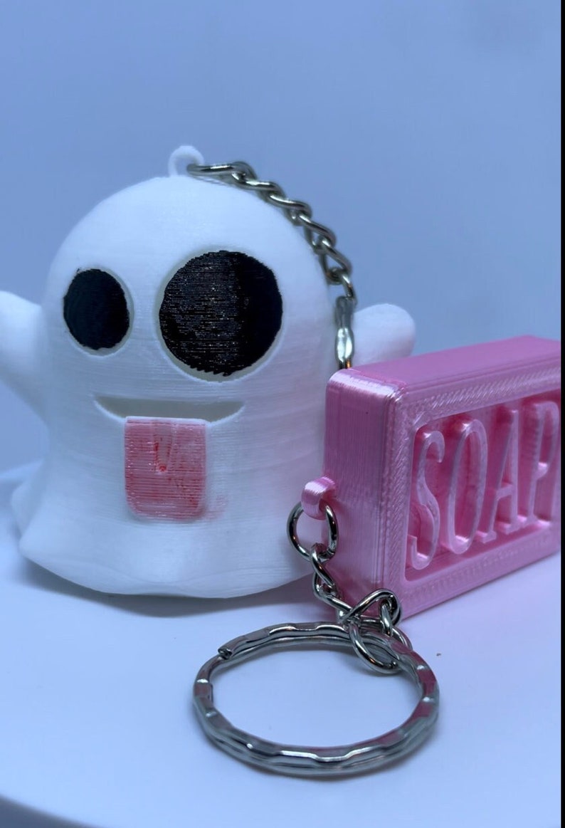 Pode incluir: Um chaveiro de fantasma branco com olhos pretos e uma l&iacute;ngua rosa, preso a um chaveiro de sabonete rosa com a palavra "SOAP" em relevo.