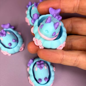 May include: A collection of small, whimsical, 3D-printed figurines. Each figurine features a light blue body with pink and purple accents. The design includes a creature-like form with a shell-like structure, perfect for crafting or collecting.