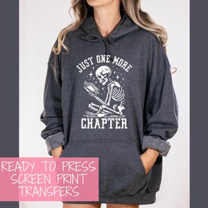 May include: Dark gray hoodie with a white graphic of a skeleton reading a book, with the text "JUST ONE MORE CHAPTER". The hoodie has a front pocket and a drawstring hood. The image also includes text that says "READY TO PRESS SCREEN PRINT TRANSFERS".