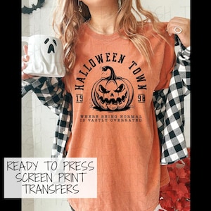May include: An orange t-shirt featuring a black graphic of a jack-o'-lantern with the text HALLOWEEN TOWN 1998 and WHERE BEING NORMAL IS VASTLY OVERRATED. The shirt is layered under a black and white plaid button-down shirt, paired with a white ghost mug.