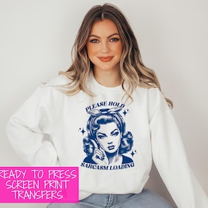 May include: White sweatshirt with a blue graphic of a woman with the text "PLEASE HOLD" above her head and "SARCASM LOADING" below. The image also includes the text "READY TO PRESS SCREEN PRINT TRANSFERS" in a pink box. The sweatshirt has long sleeves.