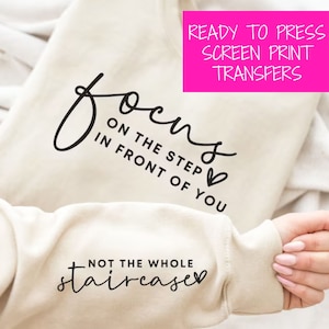 May include: A beige sweatshirt with black text that reads "Focus on the step in front of you, not the whole staircase". The text is in a cursive font and includes a small heart symbol. The sweatshirt is ready to press with a screen print transfer.
