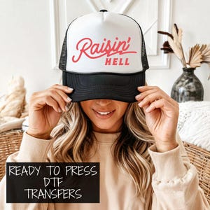 May include: A black and white trucker hat with the text "Raisin' Hell" in red script. The hat has a mesh back and a curved brim. The image also includes a black rectangle with the text "READY TO PRESS DTF TRANSFERS" in white.