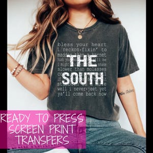以下が含まれることがあります： 暗いグレーのTシャツに、白い文字で「Bless your heart I reckon fixin' to madder than a hornet hush you I might be slower than molasses if the South well I never jeet yet ya'll come back now」と書かれた、プレス可能なスクリーン印刷転写シートです。
